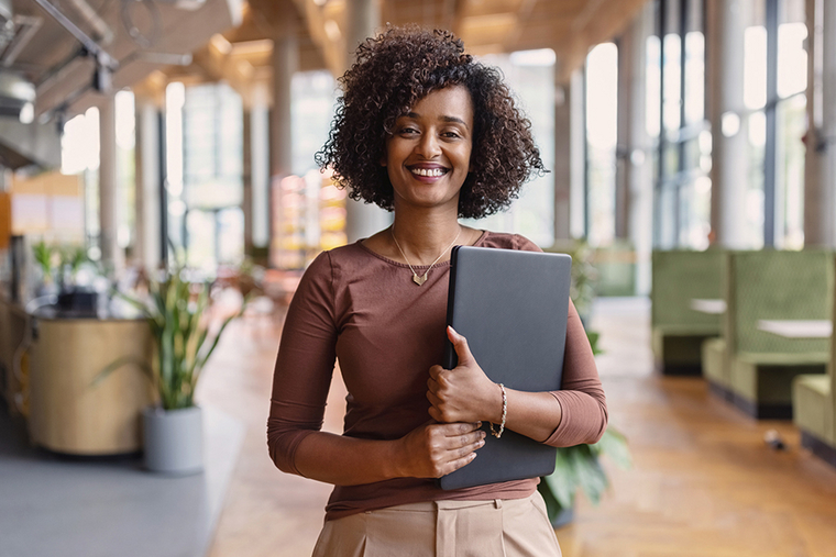 Eine Frau steht lächelnd im Büro und hält einen Laptop in der Hand 