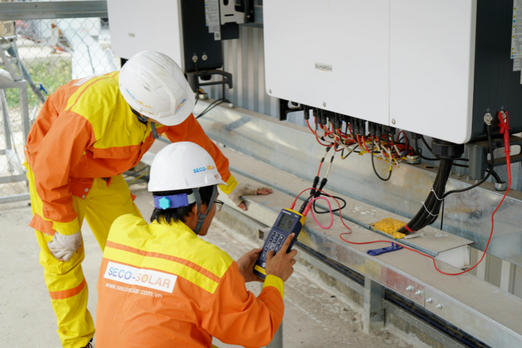 Zwei Elektroniker in Schutzkleidung und Helmen prüfen mit Messgerät die Verkabelung einer elektronischen Anlage.