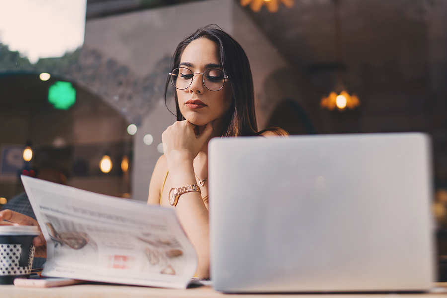 Eine Frau liest konzentriert die Zeitung und neben ihr steht ein Laptop