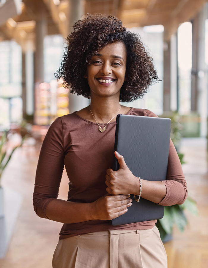 Eine Frau steht lächelnd im Büro und hält einen Laptop in der Hand