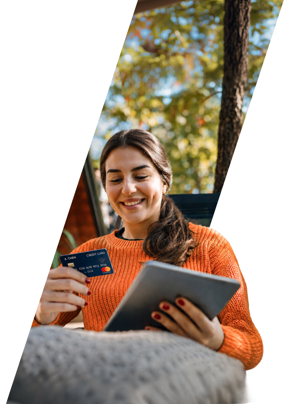 Lächelnde Frau in einem orangefarbenen Pullover hält eine Kreditkarte und ein Tablet in den Händen.