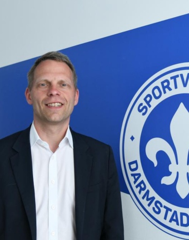 Porträt eines Mannes im dunklen Anzug vor dem Logo des SV Darmstadt 98.